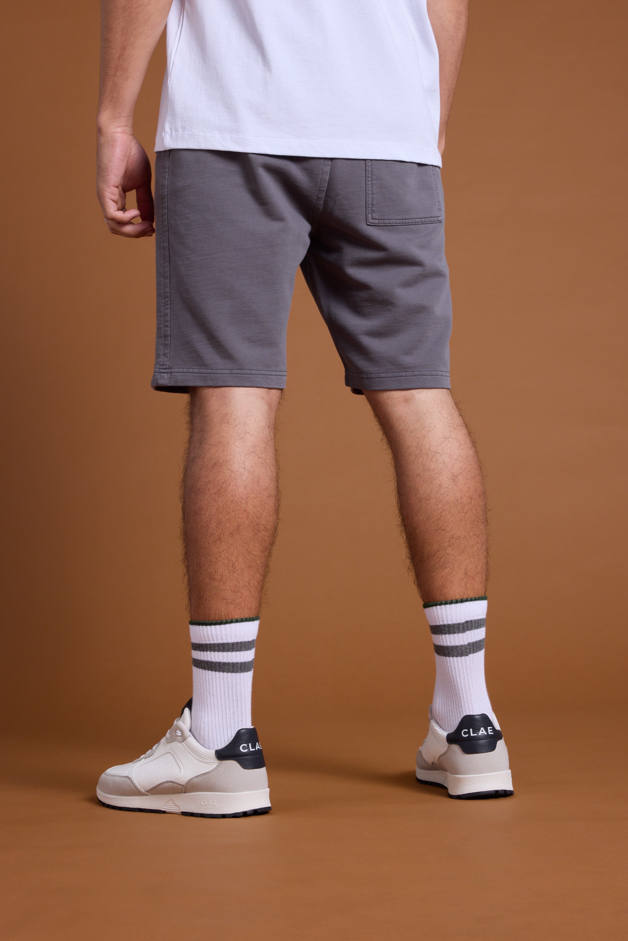 A person stands with their back to the camera, wearing Barkers Organic Classic Print Track Shorts and a white t-shirt, paired with white socks featuring black stripes and white CLAE sneakers, set against a brown background.
