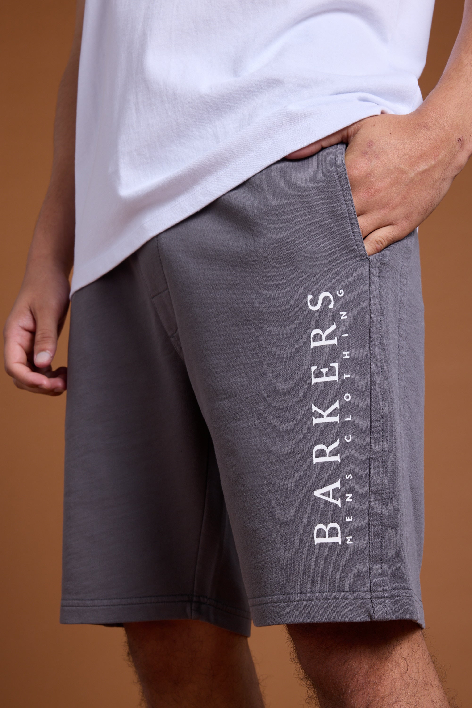 A person wears a white T-shirt and Barkers Organic Classic Print Track Shorts, featuring a relaxed fit and the brand's heritage logo with "BARKERS MENS CLOTHING" printed vertically on the gray left leg.