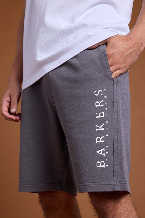 A person wears a white T-shirt and Barkers Organic Classic Print Track Shorts, featuring a relaxed fit and the brand's heritage logo with "BARKERS MENS CLOTHING" printed vertically on the gray left leg.