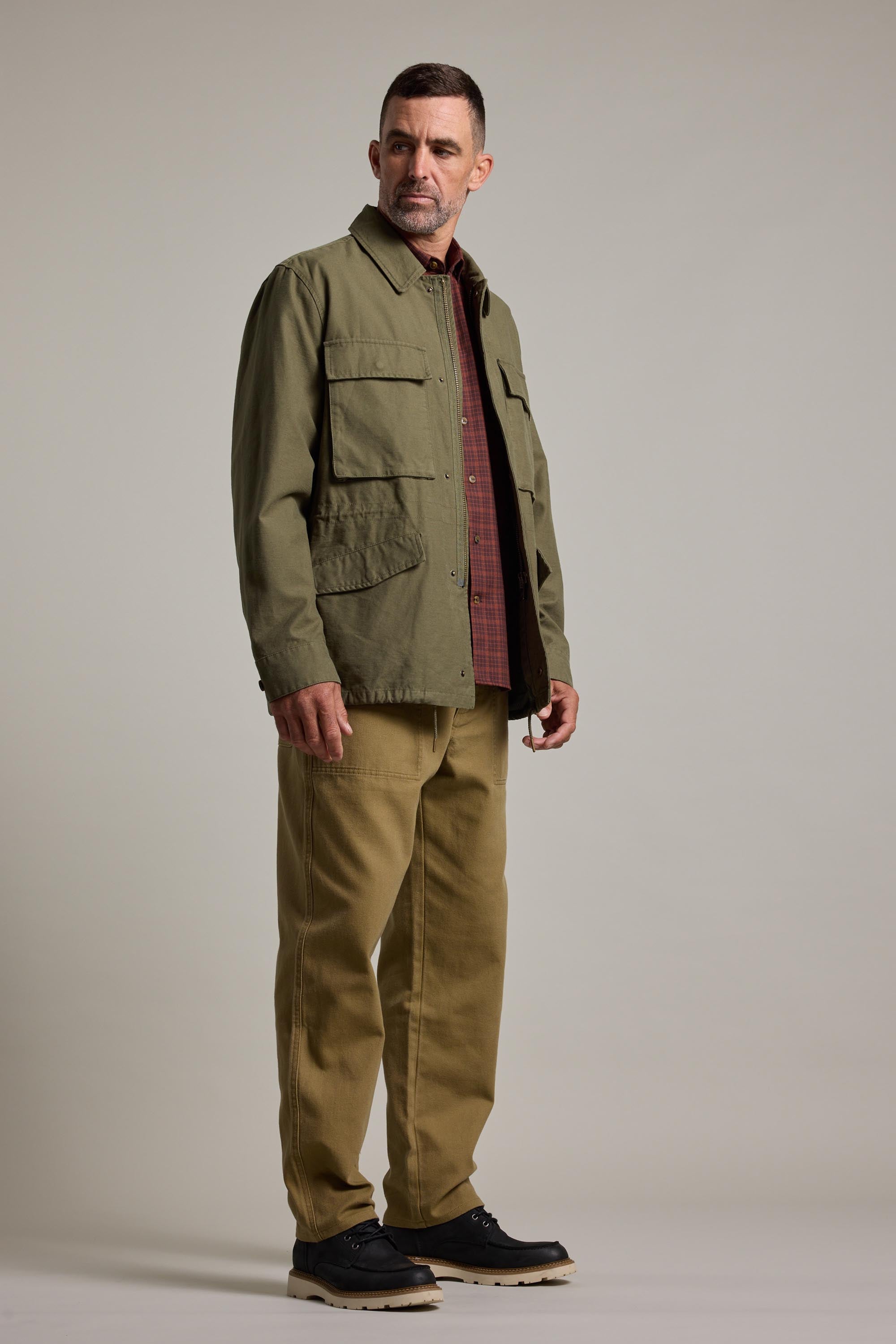 A man stands slightly to the side against a plain light background, wearing the Barkers Nowell Field Jacket—a khaki cotton utilitarian style—over a red checkered shirt, tan pants, and black shoes.