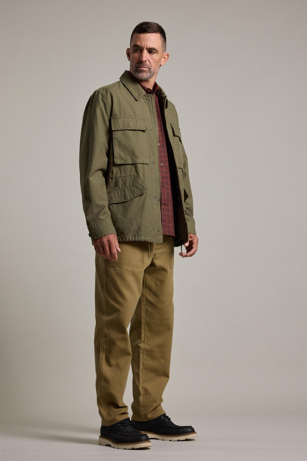 A man stands slightly to the side against a plain light background, wearing the Barkers Nowell Field Jacket—a khaki cotton utilitarian style—over a red checkered shirt, tan pants, and black shoes.