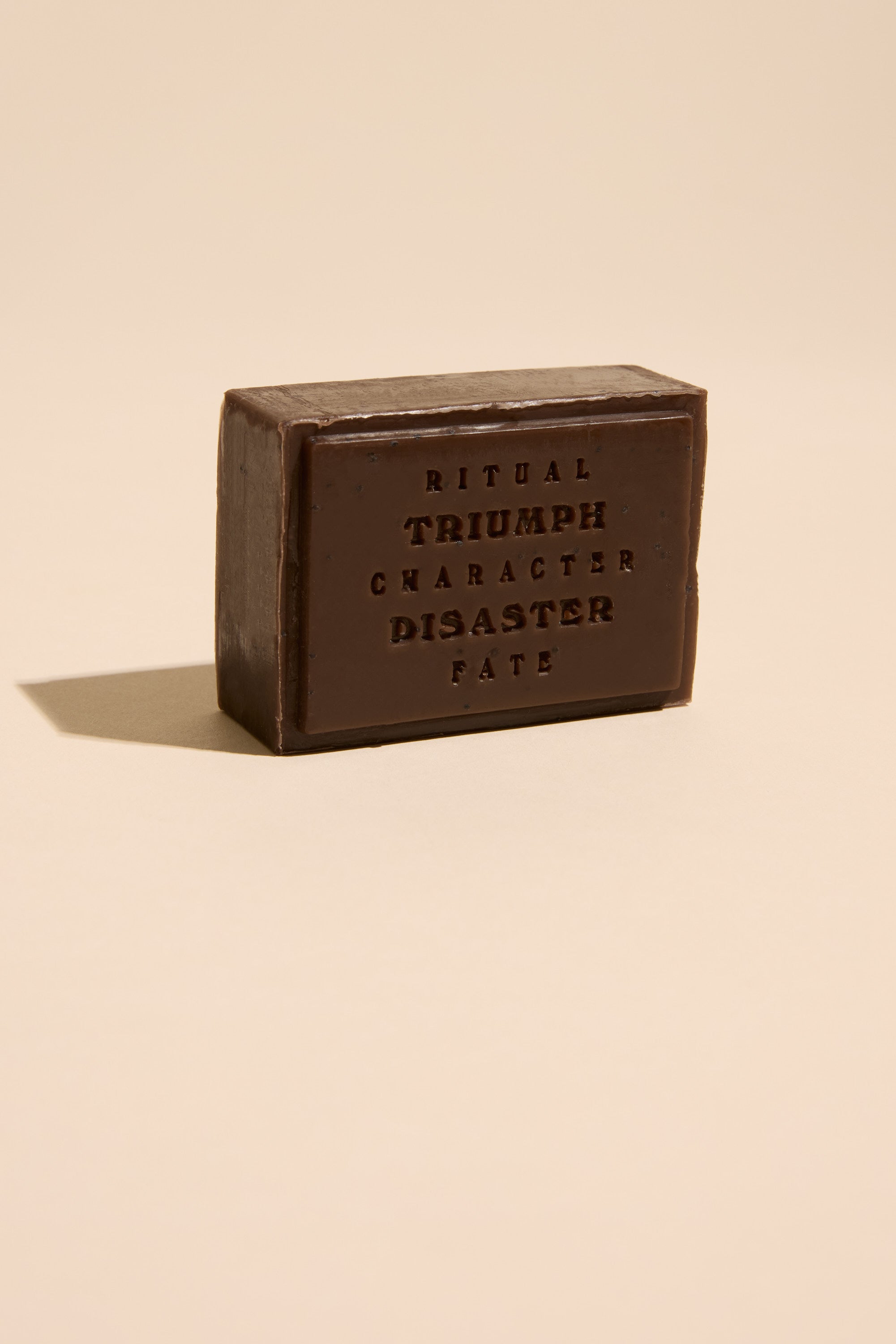 A rectangular dark brown bar of Triumph & Disaster Shearers Soap stands upright on a beige surface. Embossed with “RITUAL, TRIUMPH, CHARACTER, DISASTER, FATE,” this exfoliating soap is gentle on skin. Shadows fall to the left.