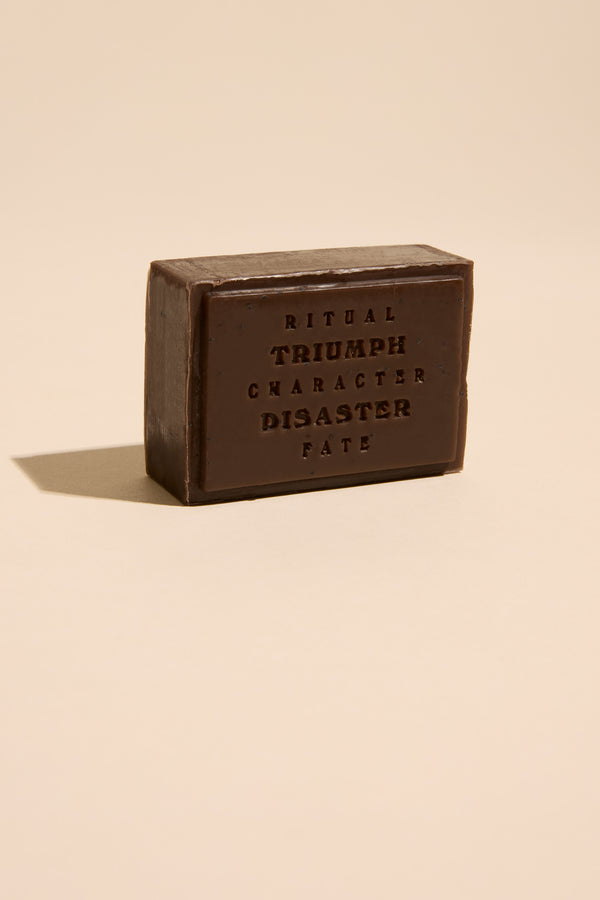 A rectangular dark brown bar of Triumph & Disaster Shearers Soap stands upright on a beige surface. Embossed with “RITUAL, TRIUMPH, CHARACTER, DISASTER, FATE,” this exfoliating soap is gentle on skin. Shadows fall to the left.