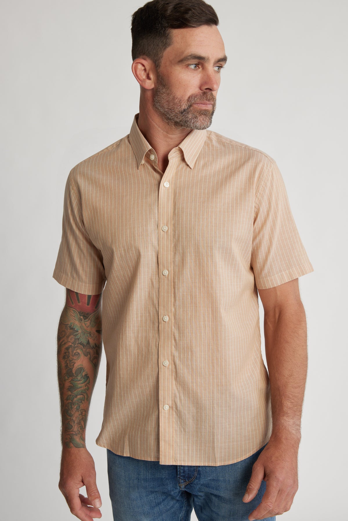 A man with short brown hair and a beard wears the Barkers Burnham Stripe Shirt and blue jeans. He has a colorful tattoo on his left forearm and looks to his left against a plain background.