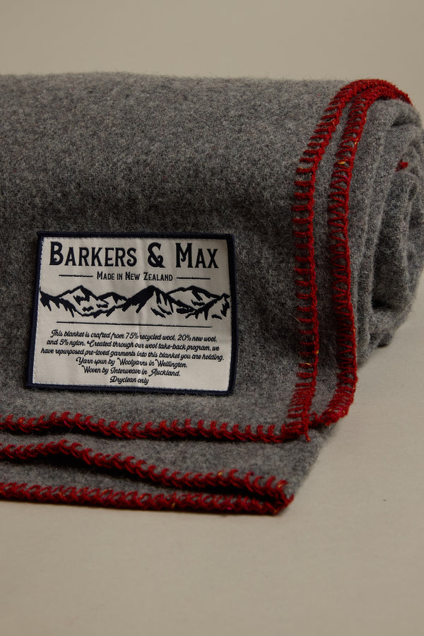A folded grey Wool Take Back Blanket by Other, made in New Zealand, features red edge stitching and a label with mountain graphic, care instructions, and text: "Barkers & Max, Made in New Zealand.