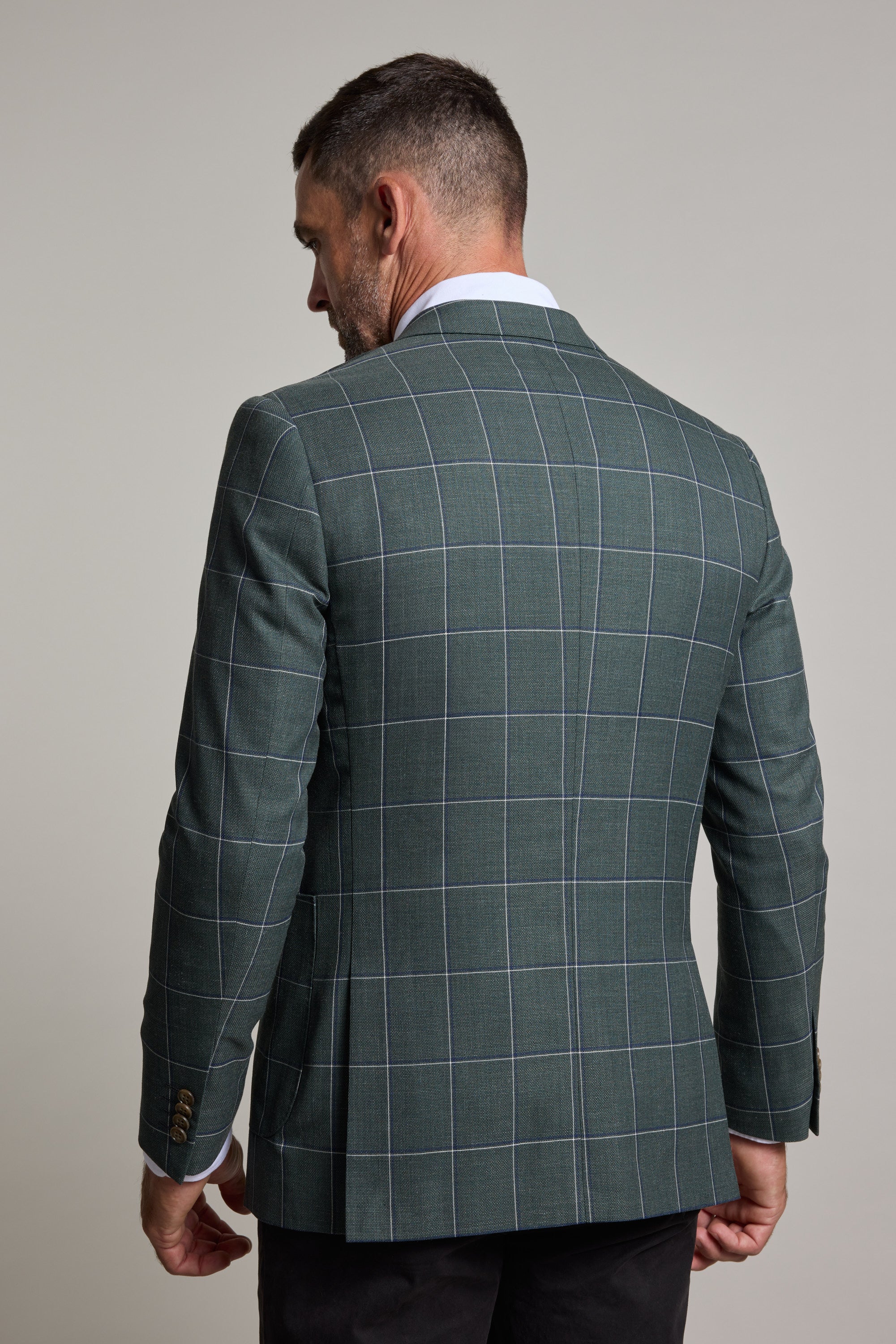 A man turned away from the camera wears a Barkers Nathaniel Check Blazer, a green wool blend men's blazer with thin white lines, styled with a white collared shirt and dark trousers, set against a plain light gray background.