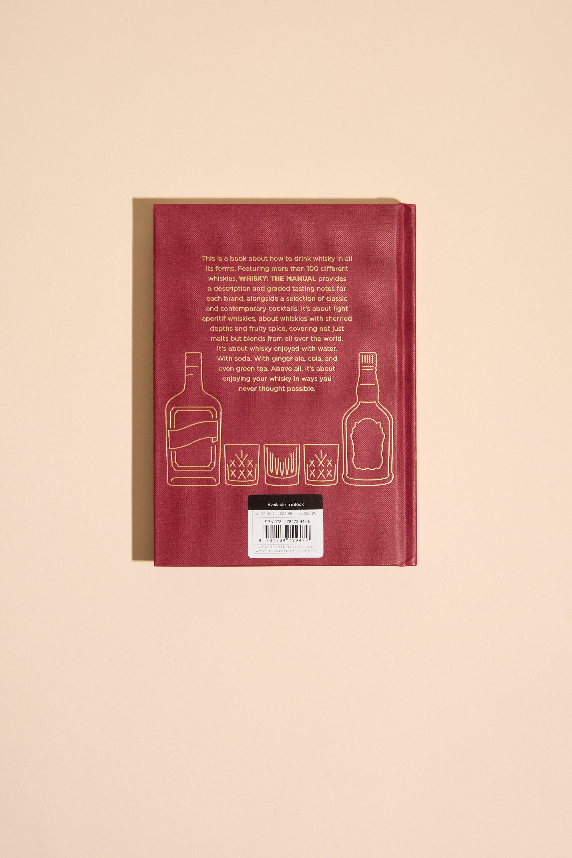 A red hardcover "Whisky The Manual" by Other lies flat, displaying its back cover with gold outlines of two liquor bottles and three whiskey glasses, plus a text block and barcode on a beige background for a whisky guide feel.