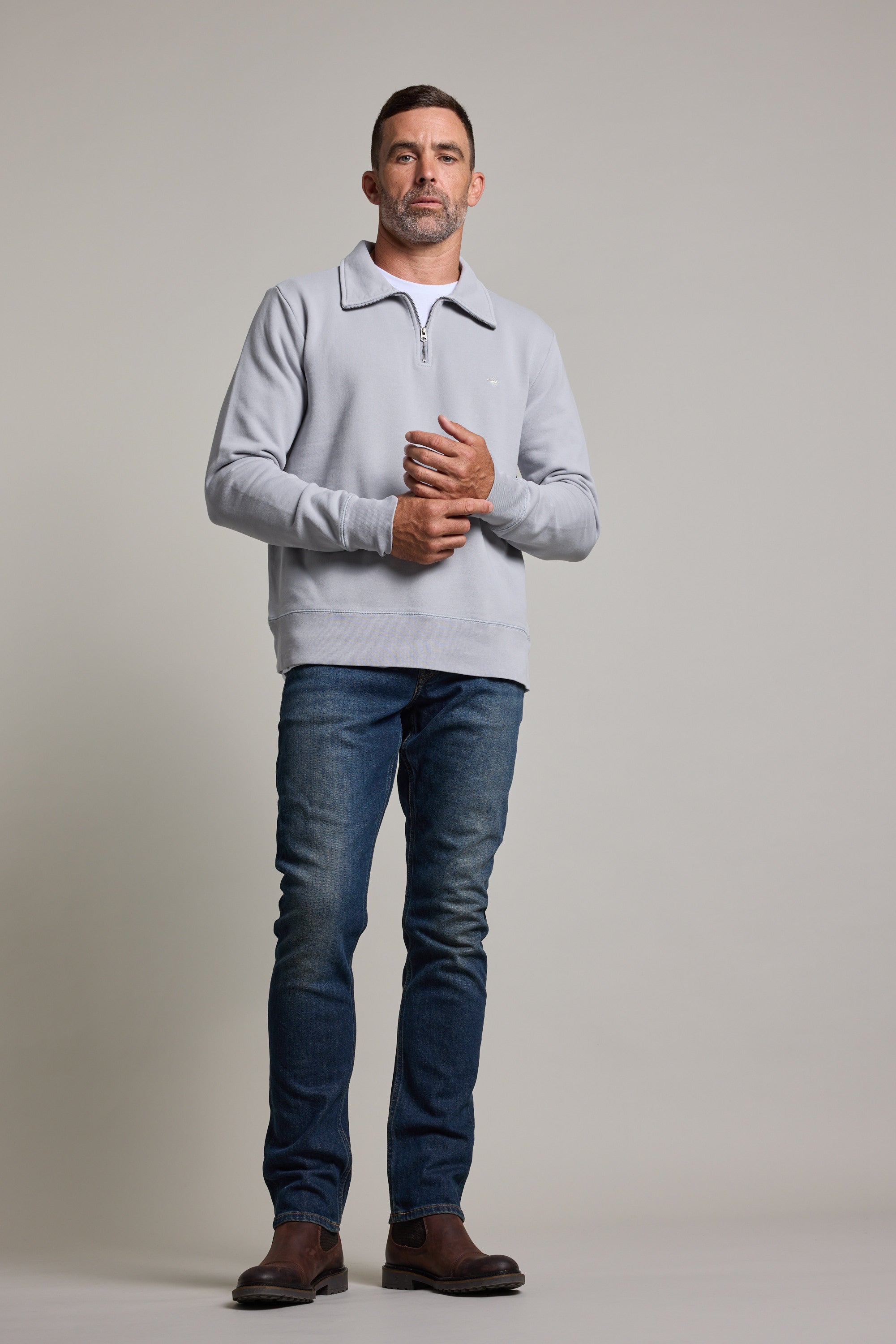 A man stands before a plain light background, wearing a Barkers Galway GD 1/4 Zip in light gray, paired with blue jeans and brown shoes. He gazes ahead with a neutral expression, hands clasped loosely in front of him.