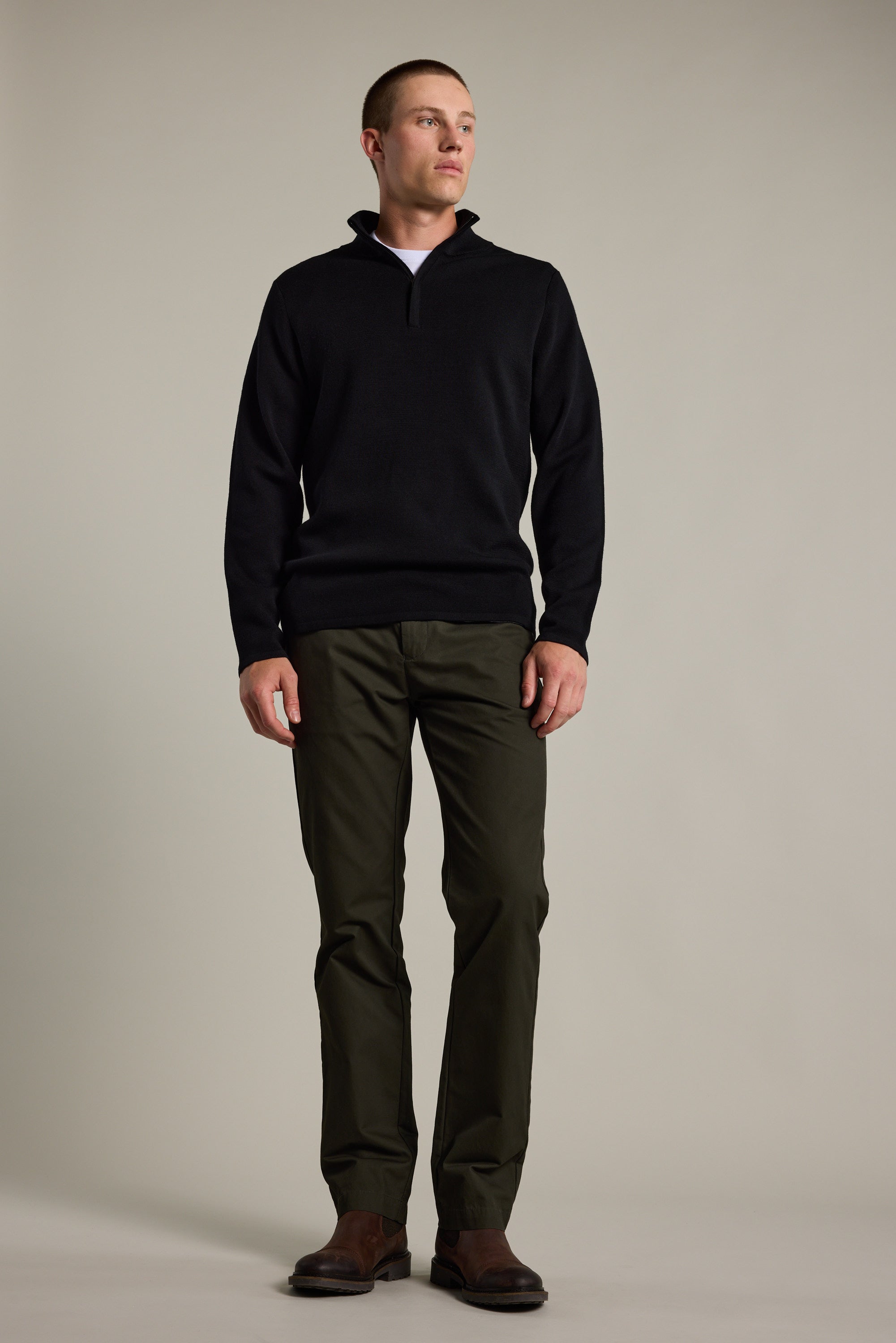 A man faces slightly to the side, wearing a black Barkers Brandon ZQRX Merino 1/4 Zip pullover, olive green pants, and brown shoes against a plain light background—ideal for cold-weather layering.