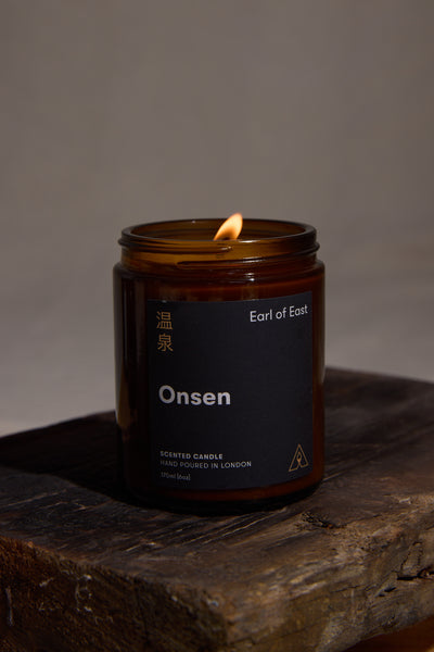 The EoE Onsen Candle 170ml by Earl of East, a peppermint-scented soy wax candle in amber glass with English and Japanese text, rests on rustic wood against a dark backdrop.