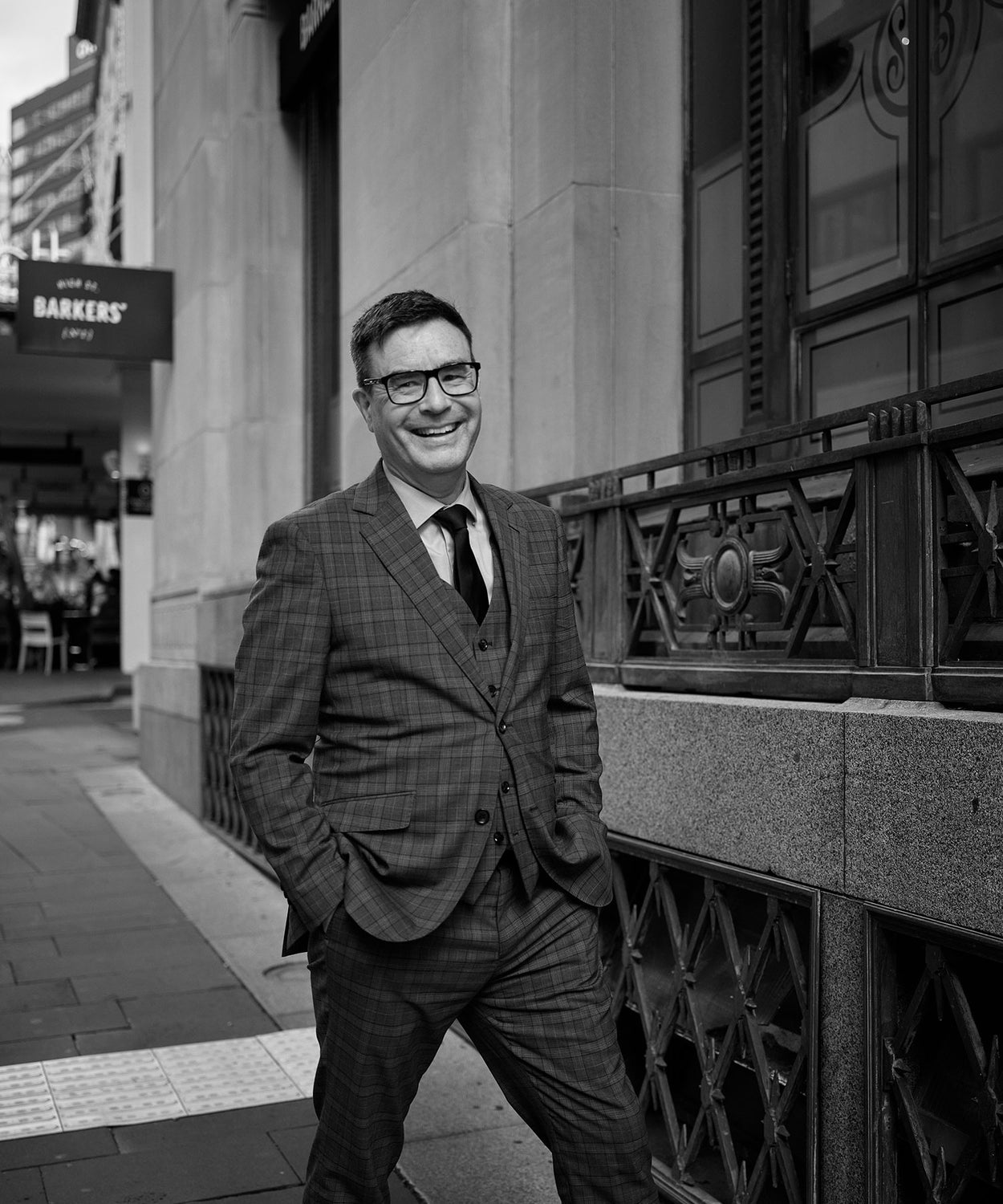 A man in a checked suit stands on a city sidewalk, smiling with his hands in his pockets. The photo is black and white, with buildings and a Barkers sign visible in the background.