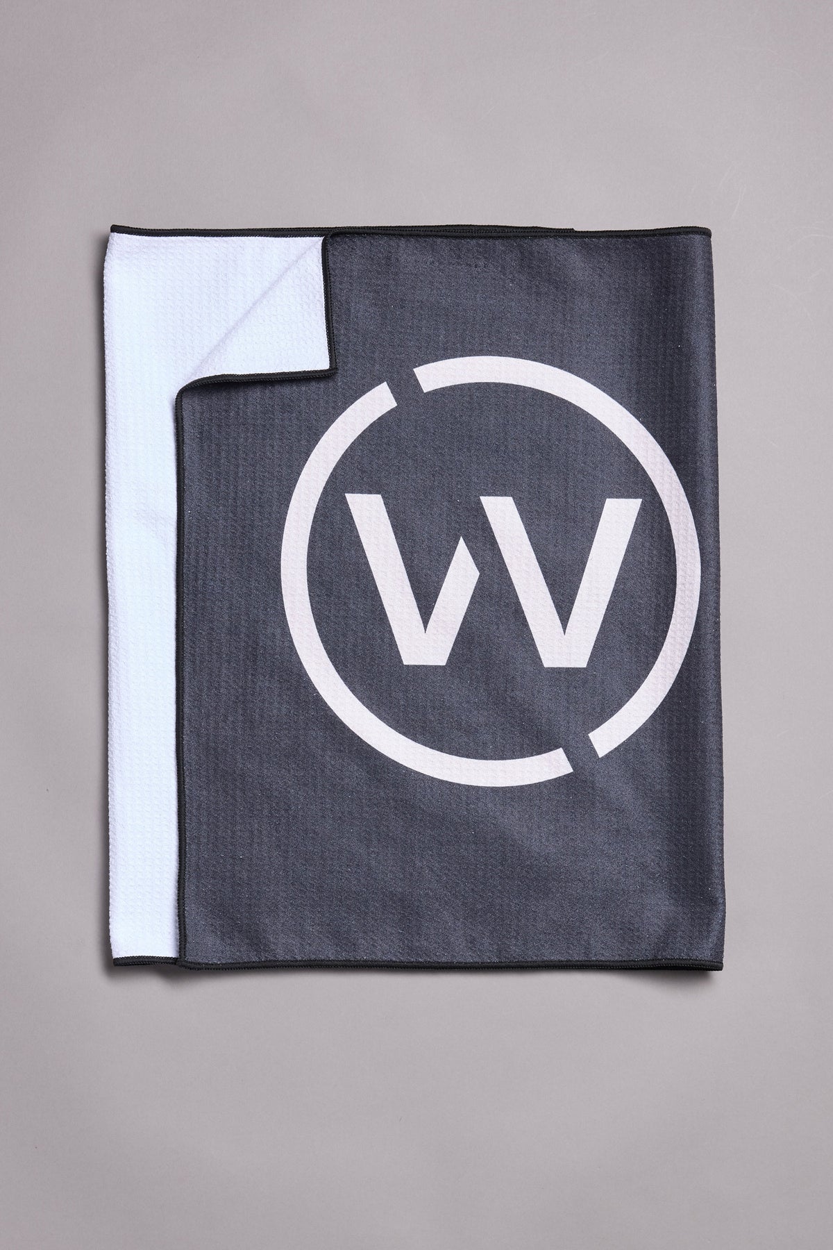 The WearEver Utility Towel by WearEver is a square black and white microfibre towel shown on a gray surface. It features a large WV logo inside a circle on the black side, with the quick-dry white inner side partially folded over a corner.