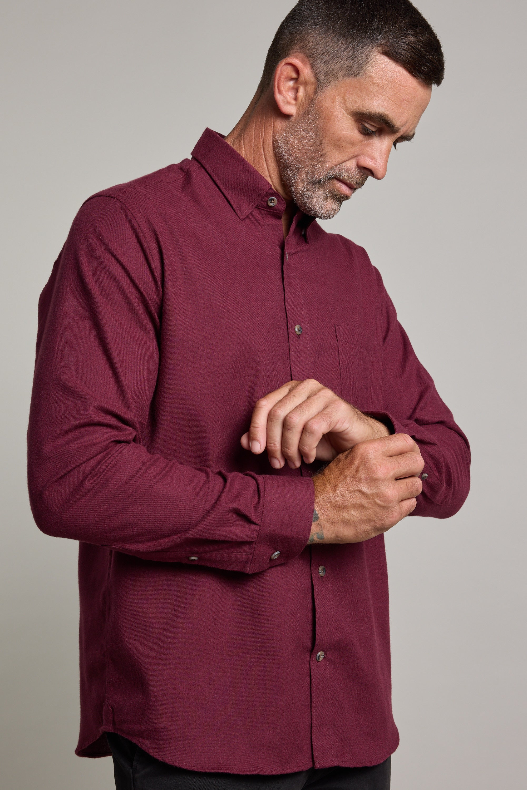 A man with short dark hair and a beard wears the Barkers Antonio Shirt, a classic fit, long-sleeved maroon style in a cotton-wool blend. He looks down while fastening his cuff against a neutral background.