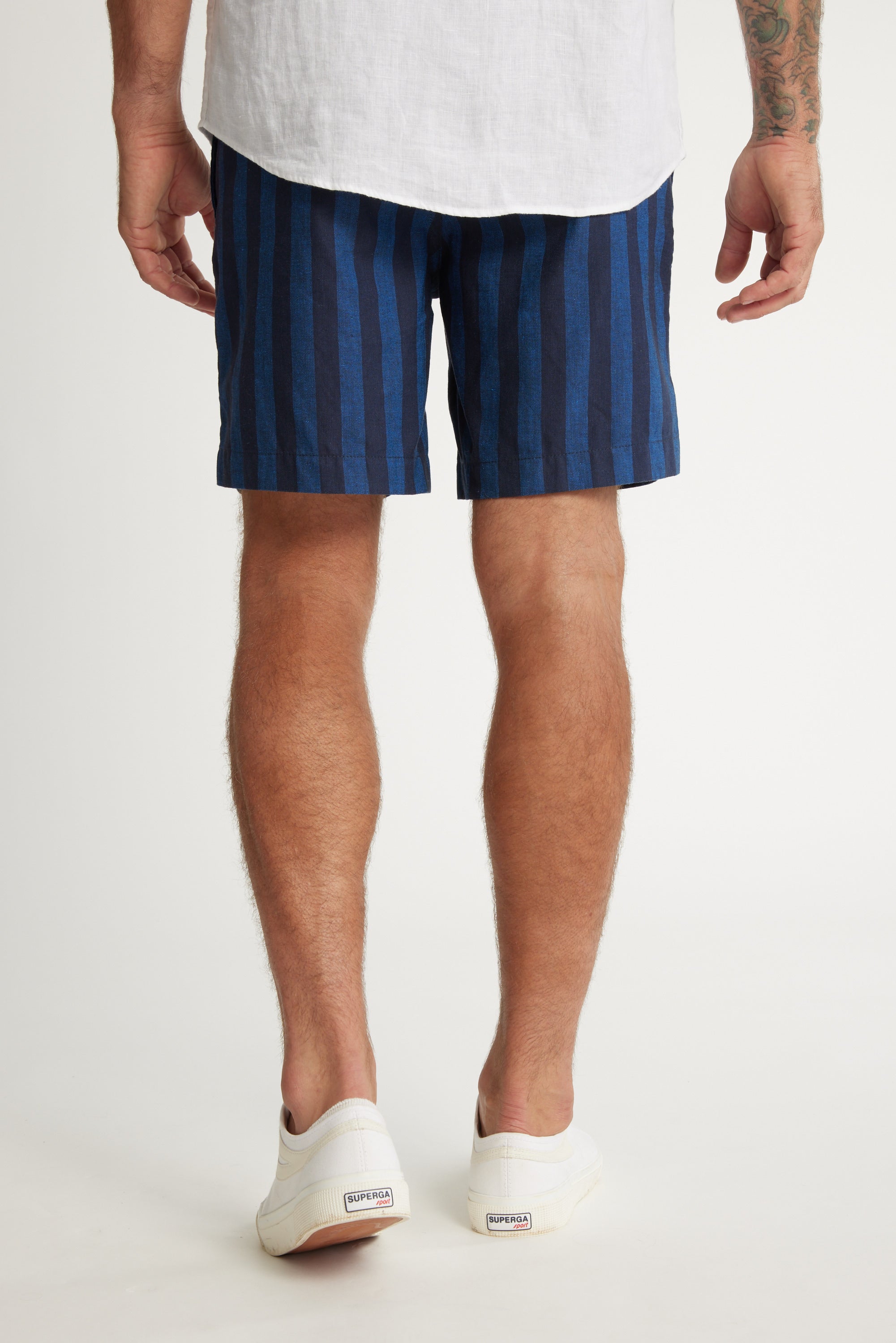 A man stands with his back to the camera wearing a white shirt, Barkers Ashbury Stripe Short in blue and navy, and white slip-on shoes. His arms hang by his sides, revealing part of a tattoo on his left forearm.