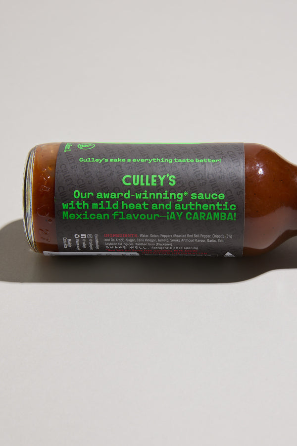 A bottle of Culley’s Cheeky Chipotle Hot Sauce rests on its side, showing a label that reads, “Our award-winning sauce with mild heat and authentic Mexican flavour – ¡AY CARAMBA!” Roasted red bell pepper is highlighted in the ingredients below.