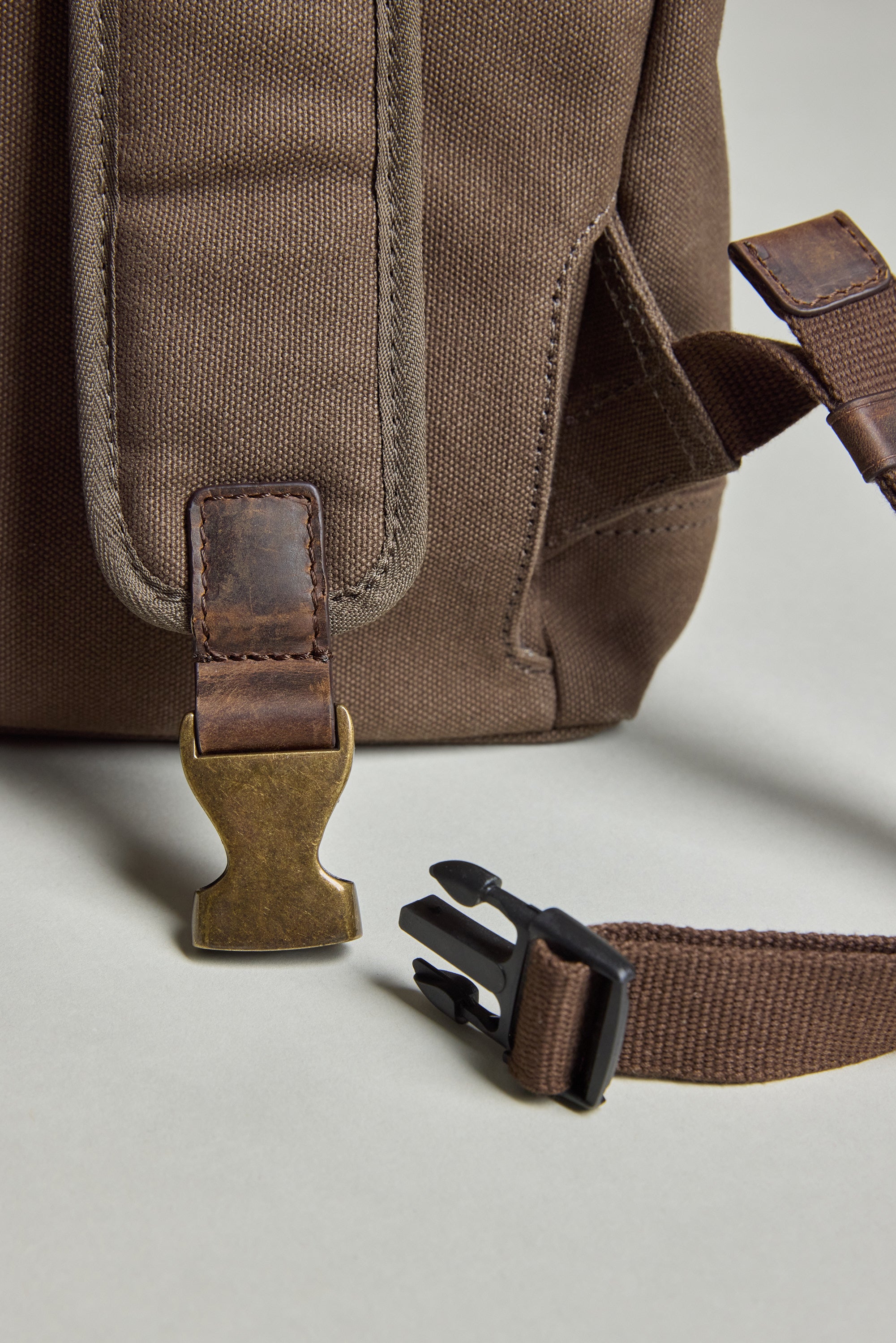 Close-up of Barkers’ Foxton Canvas Convertible Tote in brown cotton canvas with a leather strap and brass buckle, shown beside a dark brown nylon strap—highlighting its 3-in-1 versatility, all resting on a light surface.