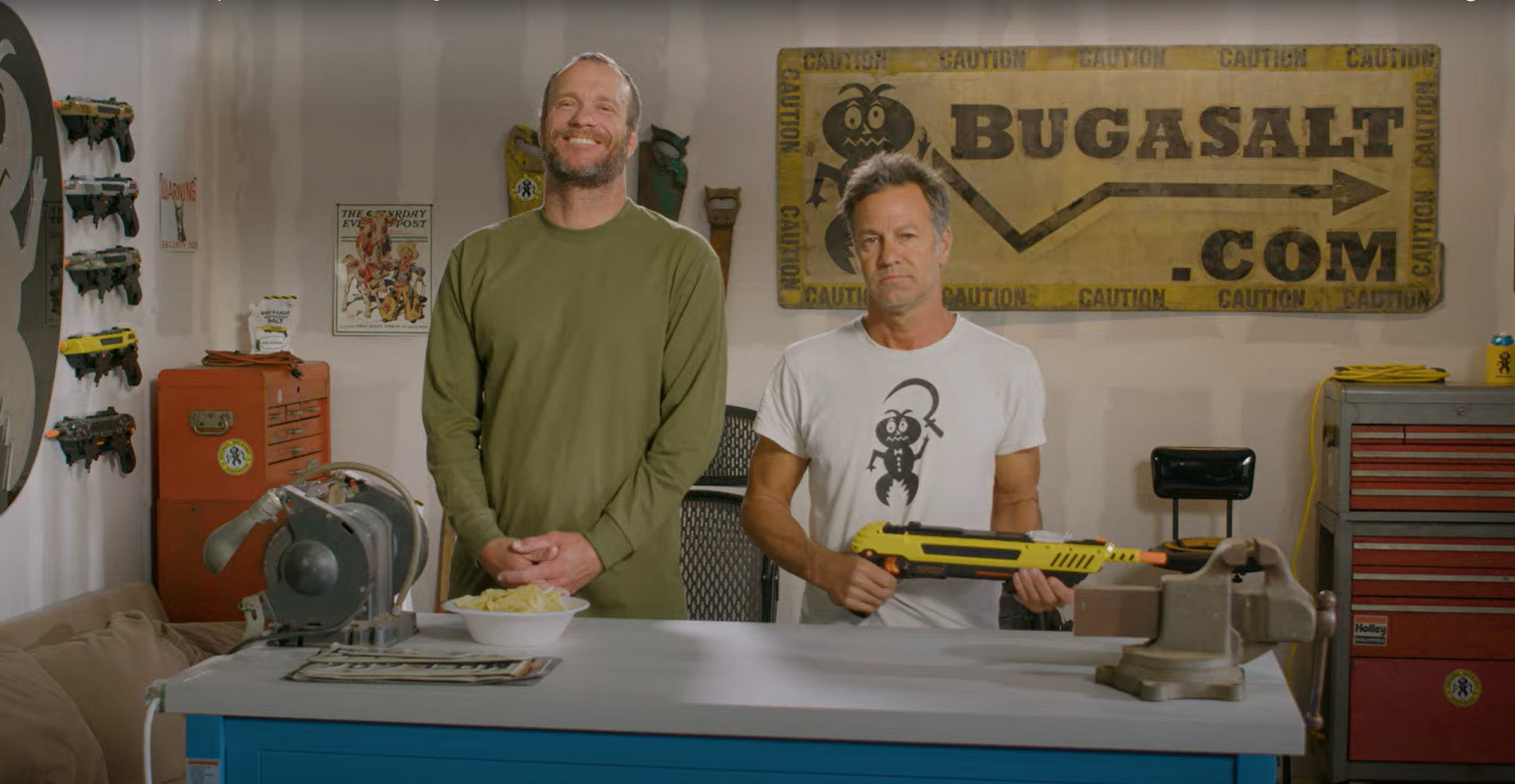 Two men stand in a workshop; one smiles with hands clasped, the other holds a yellow Bug-A-Salt gun. Behind them is a large sign reading BUGASALT.COM and various tools and decorations.