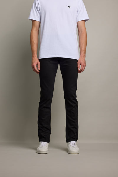 A person, head not visible, stands against a plain background wearing a white t-shirt, Barkers Rambler Denim Slim Jean, and white sneakers.