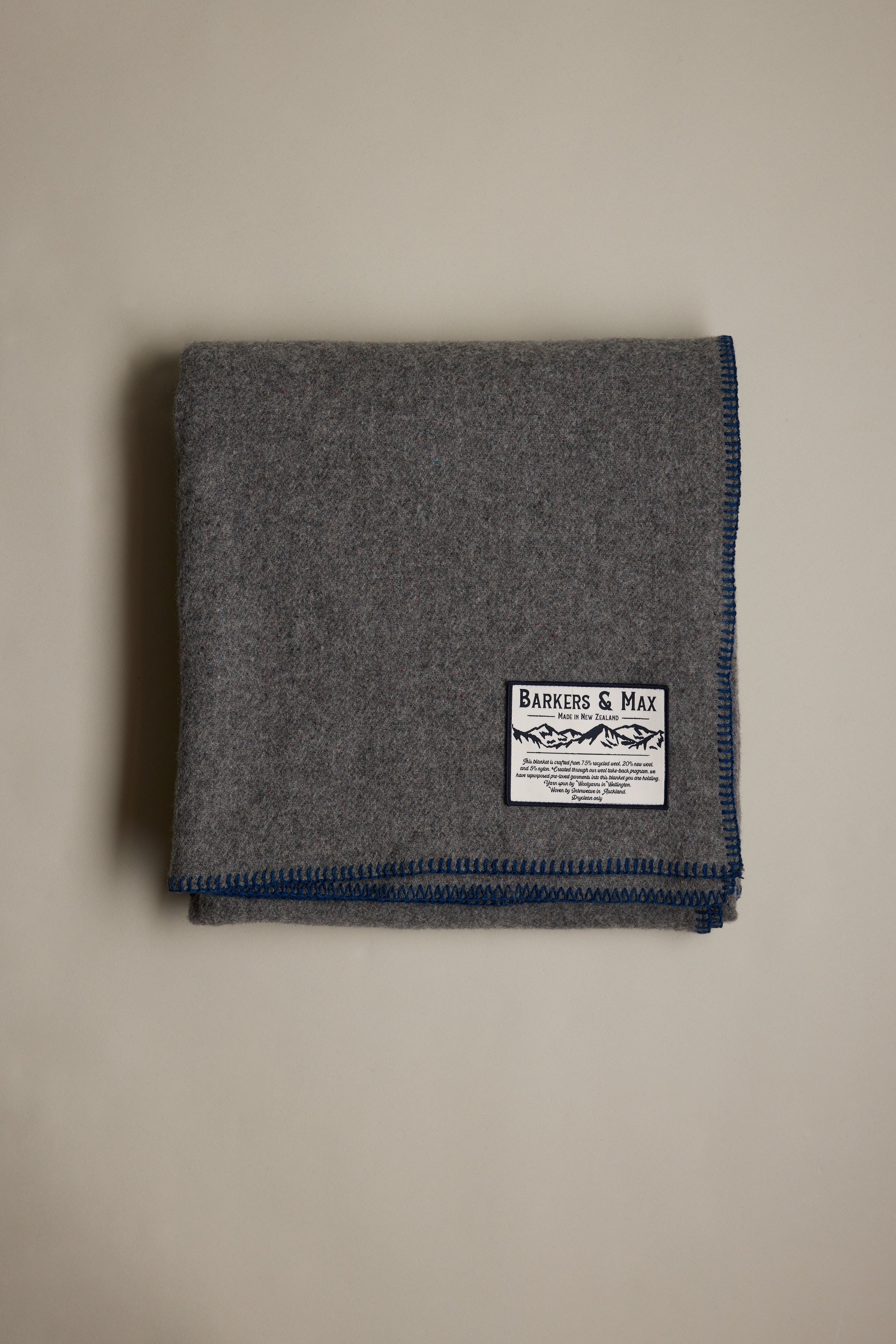 The Wool Take Back Blanket by Other, made from sustainable gray wool with blue-stitched edges and a white Barkers & Max label, is shown folded on a plain, light-colored surface.