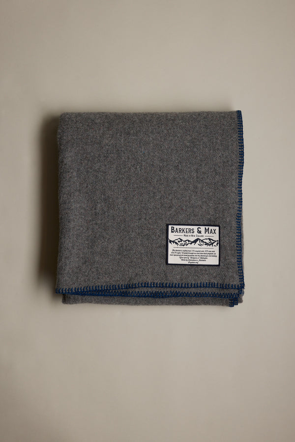The Wool Take Back Blanket by Other, made from sustainable gray wool with blue-stitched edges and a white Barkers & Max label, is shown folded on a plain, light-colored surface.