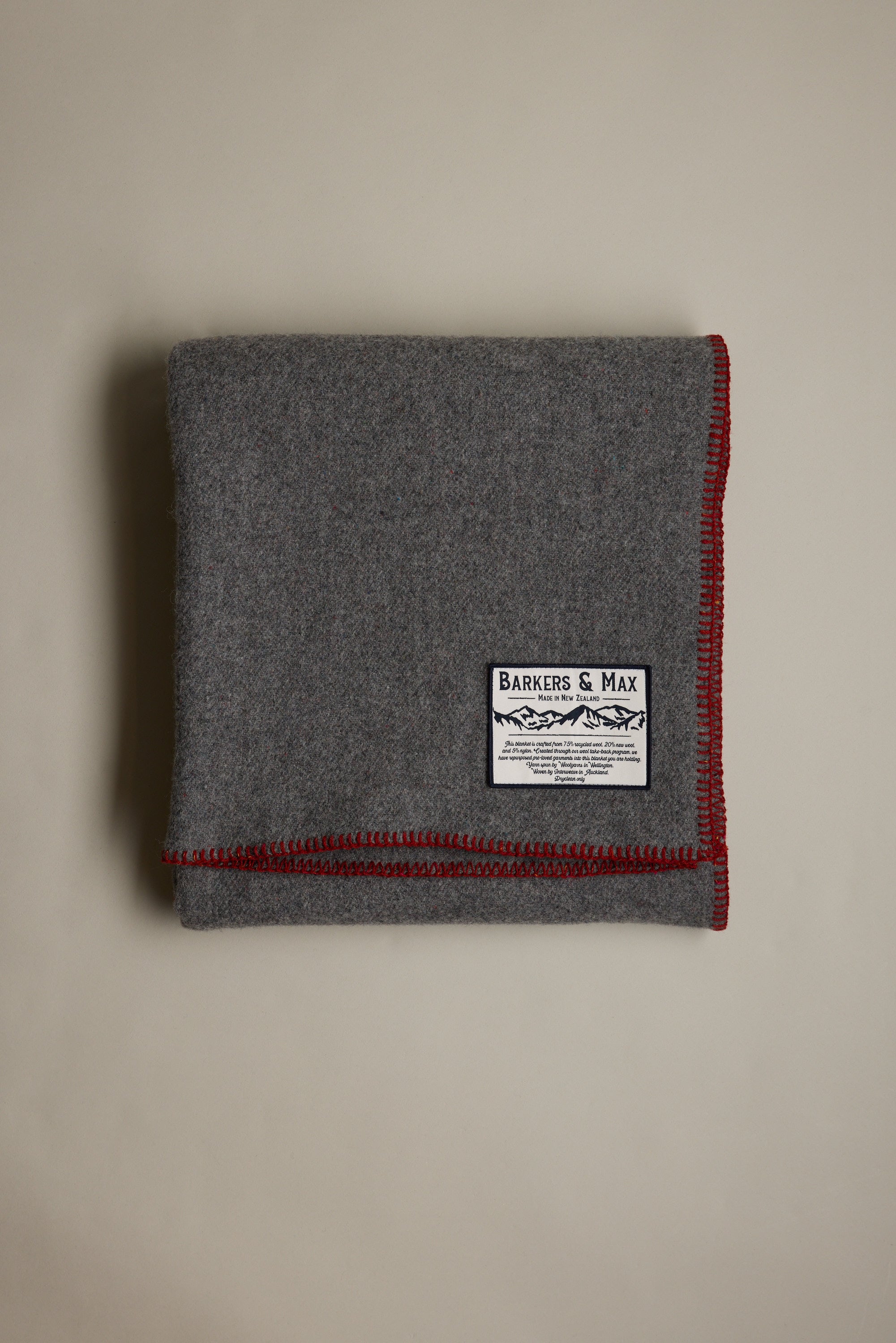A folded gray Wool Take Back Blanket by Other, made from recycled wool, with red-stitched edges and a white Barkers & Max label, displayed on a plain light background.