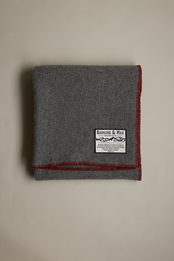 A folded gray Wool Take Back Blanket by Other, made from recycled wool, with red-stitched edges and a white Barkers & Max label, displayed on a plain light background.