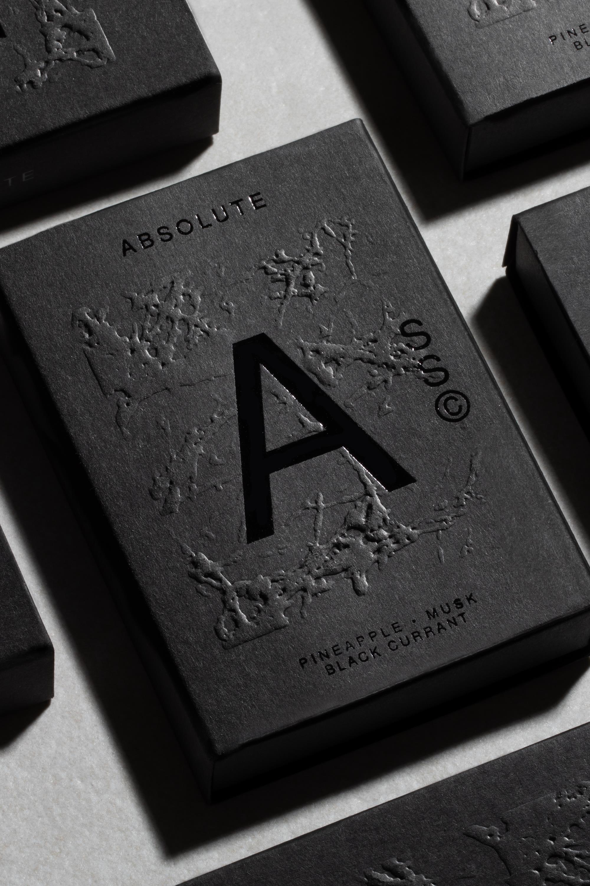 A close-up of Solid State - Absolute, a pocket-sized black box cologne embossed with an "A," textured botanical motifs, and the notes "Pineapple, Musk, Black Currant." Creed Aventus-inspired scent by Solid State; other boxes surround it.