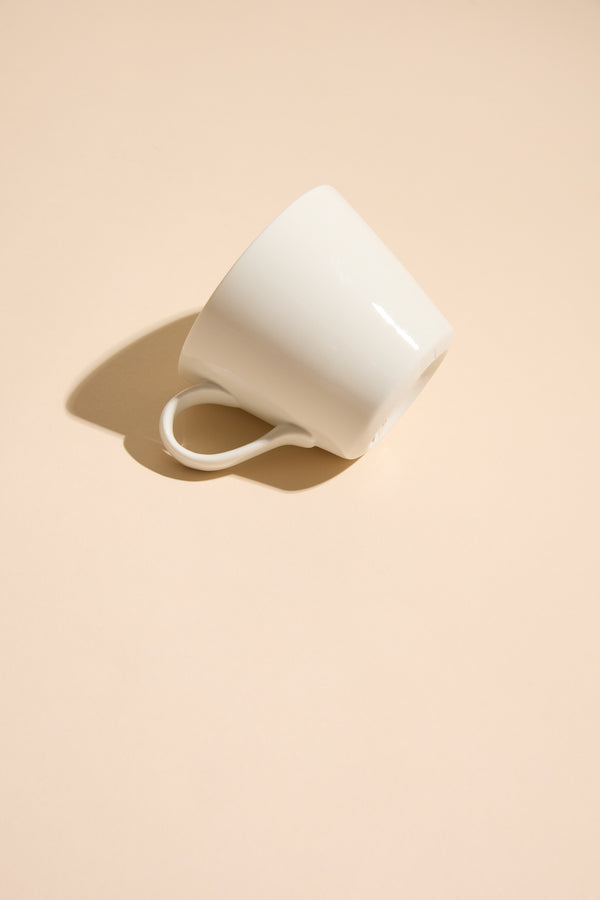 A white "Bucket Mug - Grumpy" by Other lies on its side on a light beige surface, casting a soft shadow in a minimalistic and clean composition.