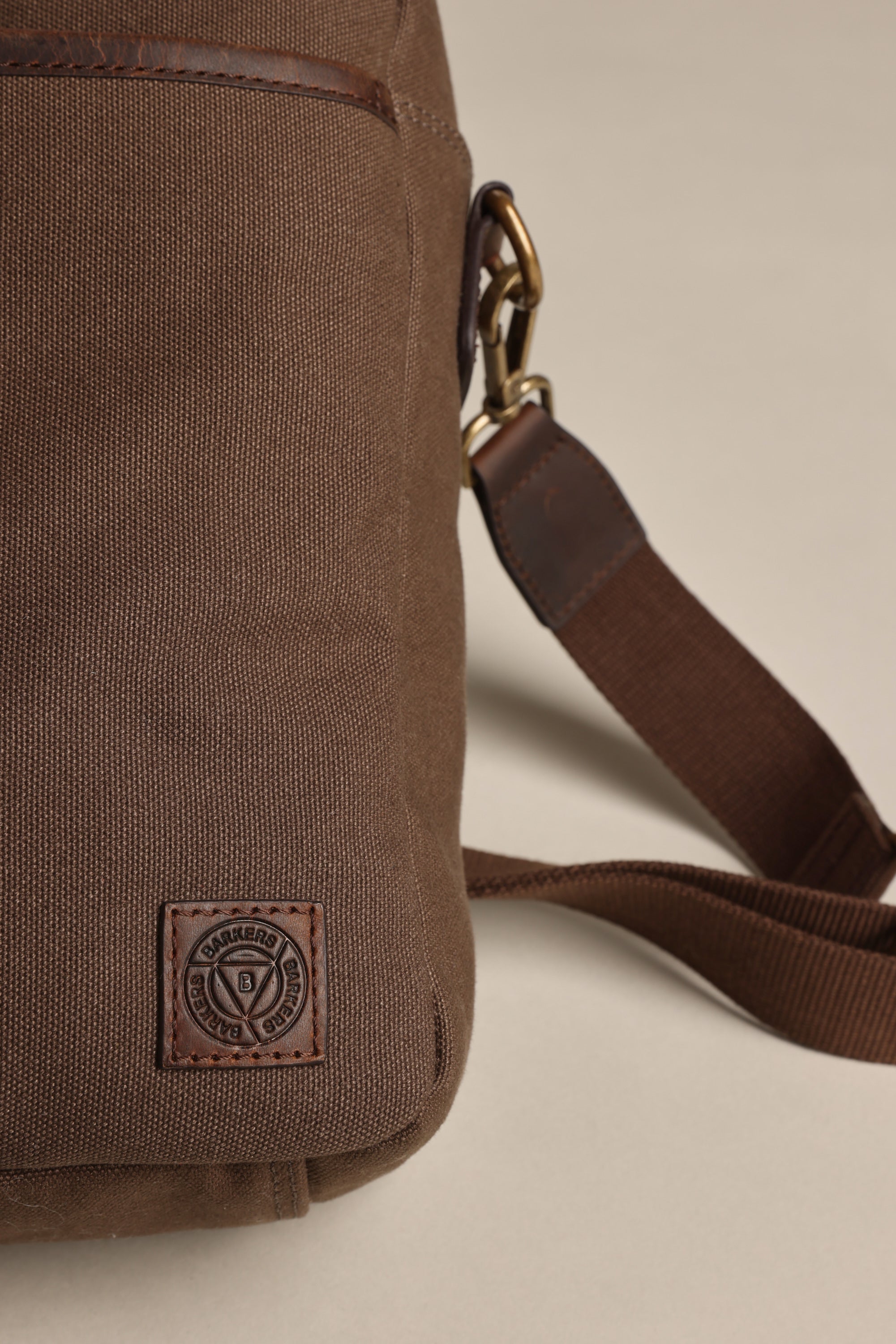 A close-up of the Barkers Redford Canvas Laptop Bag, featuring a premium leather patch with a circular logo and a brown adjustable shoulder strap with brass clasp, set against a light beige background.