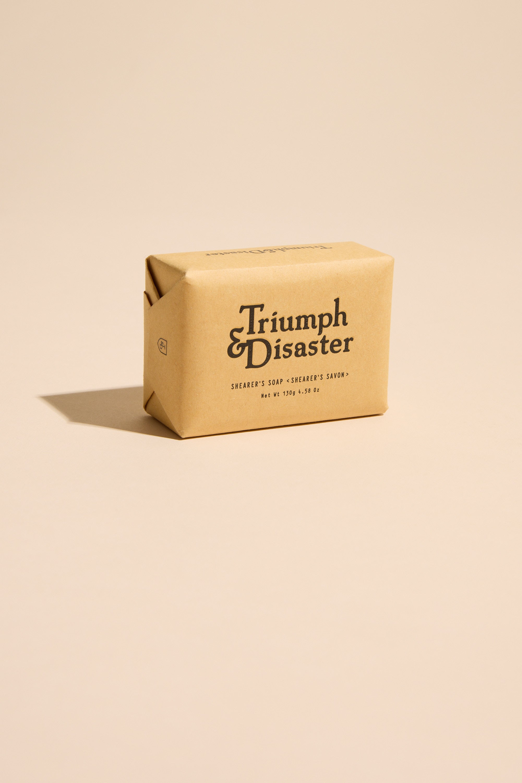A bar of Triumph & Disaster Shearers Soap, wrapped in branded brown paper, rests on a beige surface with a soft shadow. This exfoliating soap gently cares for skin while cleansing, seamlessly blending care and efficacy.