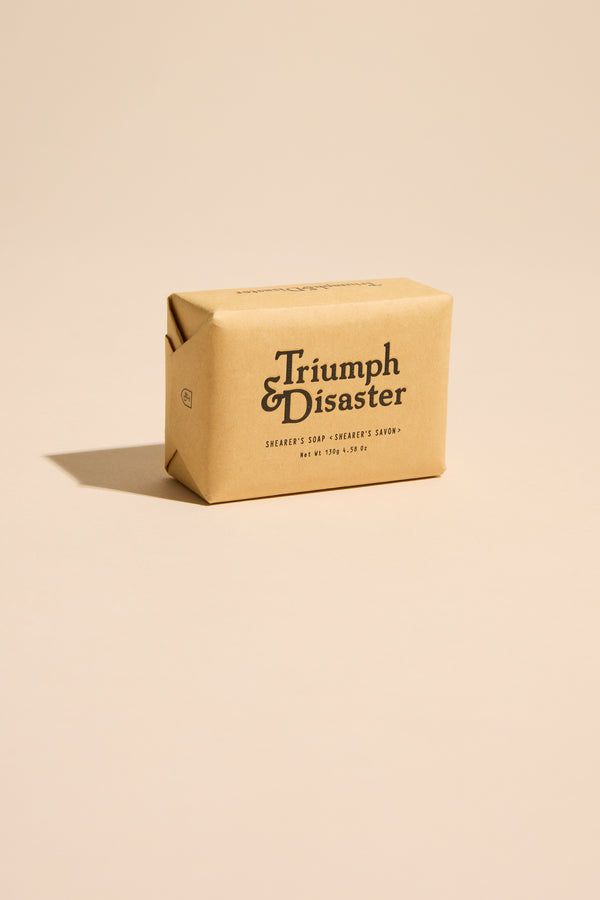 A bar of Triumph & Disaster Shearers Soap, wrapped in branded brown paper, rests on a beige surface with a soft shadow. This exfoliating soap gently cares for skin while cleansing, seamlessly blending care and efficacy.
