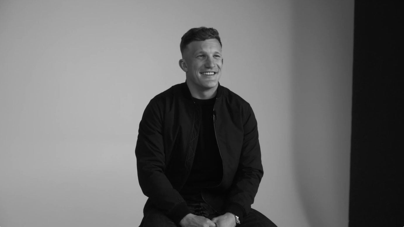 Man sitting and smiling in a studio with a plain, light NZ background.