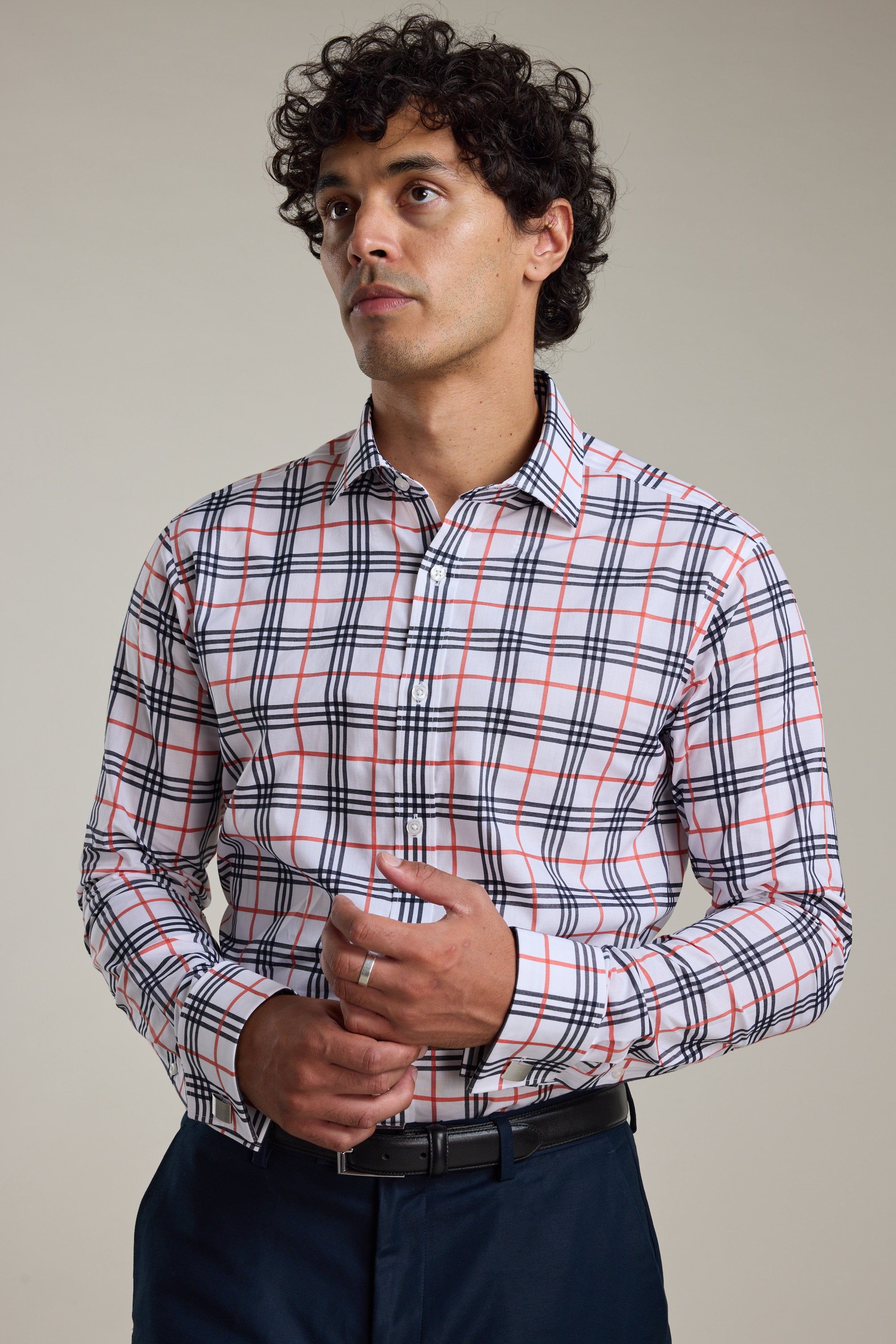A man with curly hair wears the Barkers Wilma French Cuff Business Shirt, featuring a red, black, and white plaid pattern, with dark pants. He stands against a plain background, looking slightly upward with his hands together.