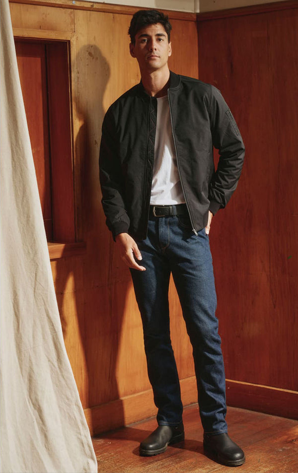 Man in black jacket, white shirt, and boots stands in a NZ wood-paneled room.
