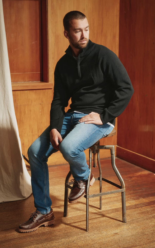 Man in a black sweater and jeans sits on a stool in a wooden NZ room, looking to the side.
