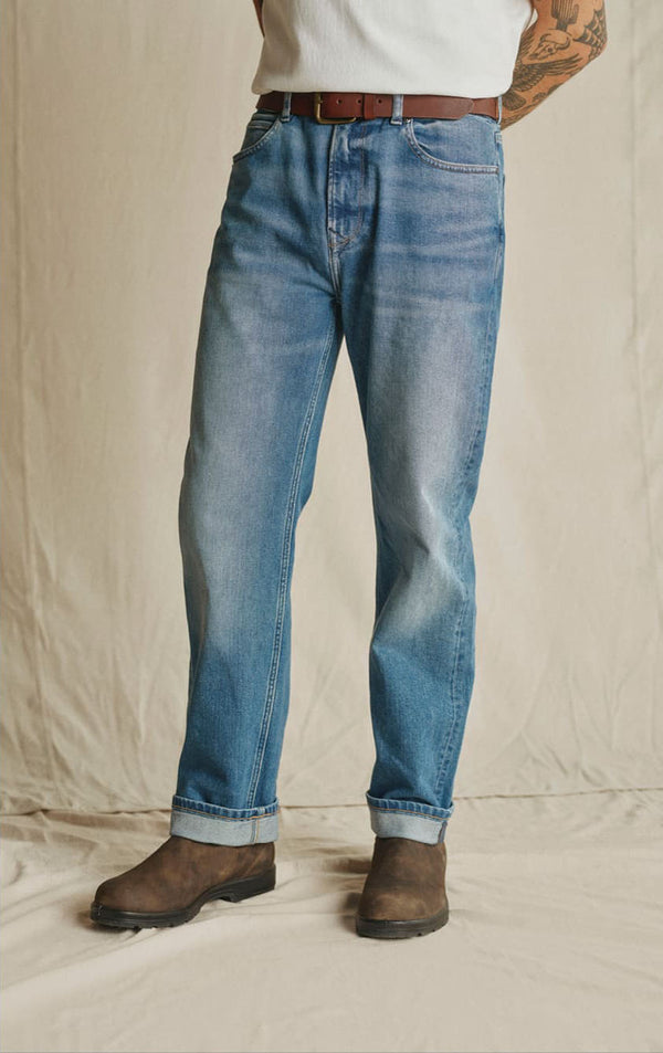Person wearing blue jeans, brown boots, and a white shirt, standing on an NZ-inspired backdrop.