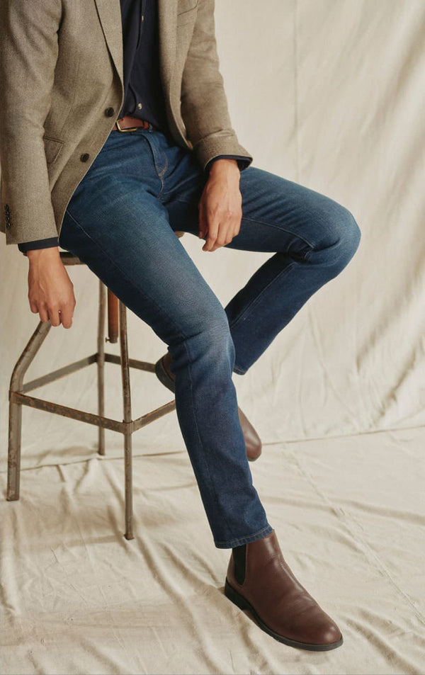 Man sitting on a stool, wearing a blazer, blue jeans, and brown boots on an NZ-inspired backdrop.
