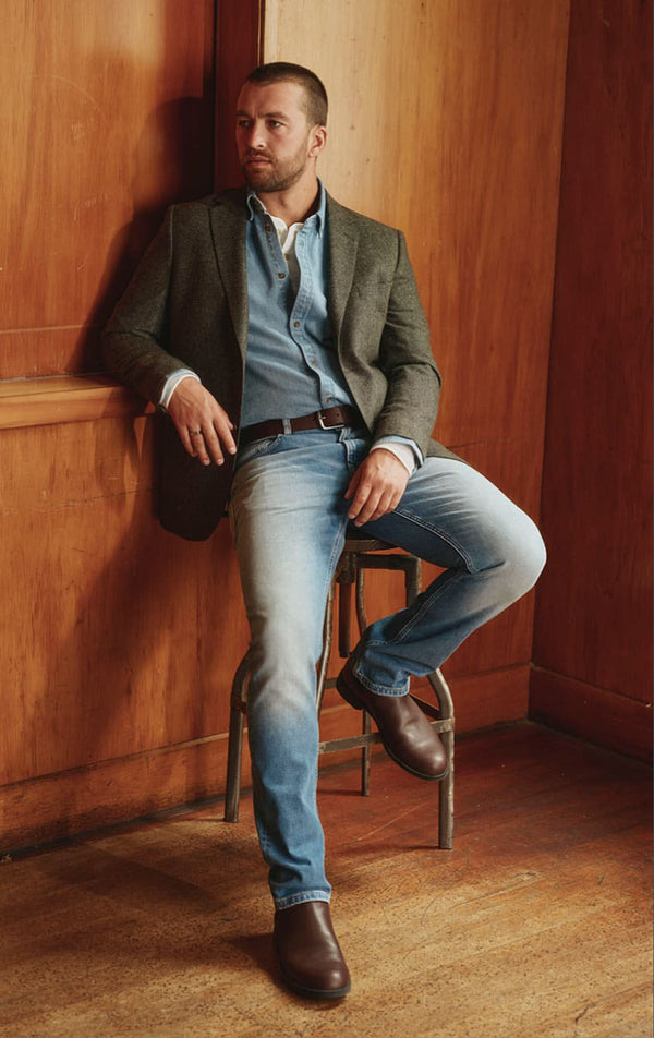 Man in a blazer, denim shirt, and jeans sits on a stool in a NZ wood-paneled room.