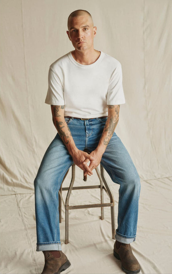 Man with tattoos in white shirt and blue jeans sitting on a stool, styled in a cool NZ vibe.