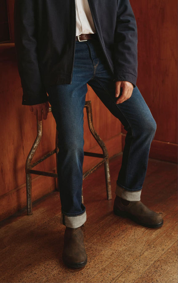 Person wearing cuffed jeans, brown boots, and a dark jacket sits on a metal NZ stool.
