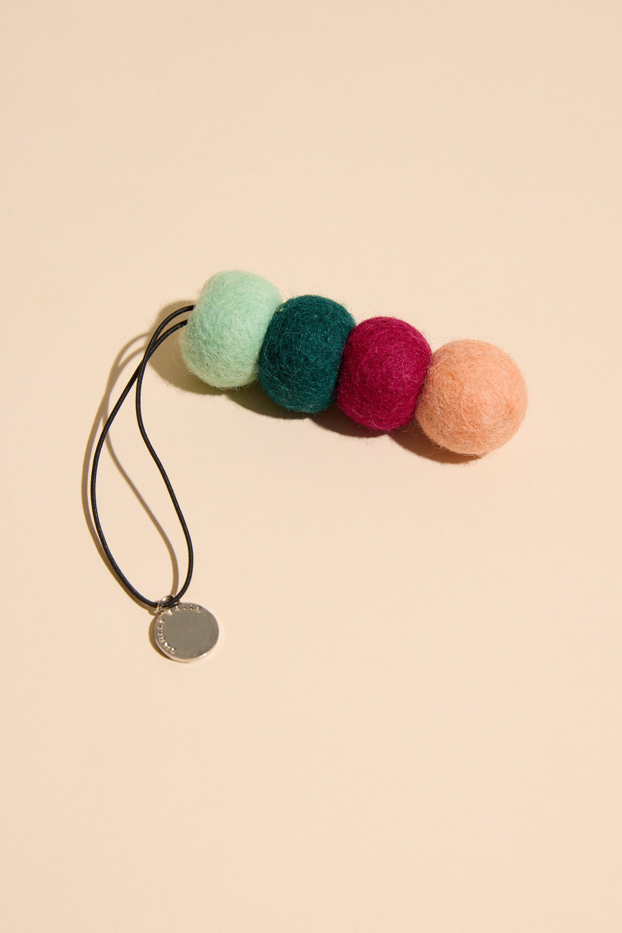 A black elastic hair tie with four large felt balls in pastel mint, dark green, burgundy, and peach plus a metallic charm—evoking the Smelly Balls: Roadie / Tobacco Vanilla style by Other.