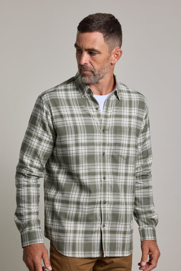 A man with short brown hair and a beard wears a green check Barkers Paxtonheck Shirt over a white t-shirt and tan pants, standing against a plain light background, looking to his left.