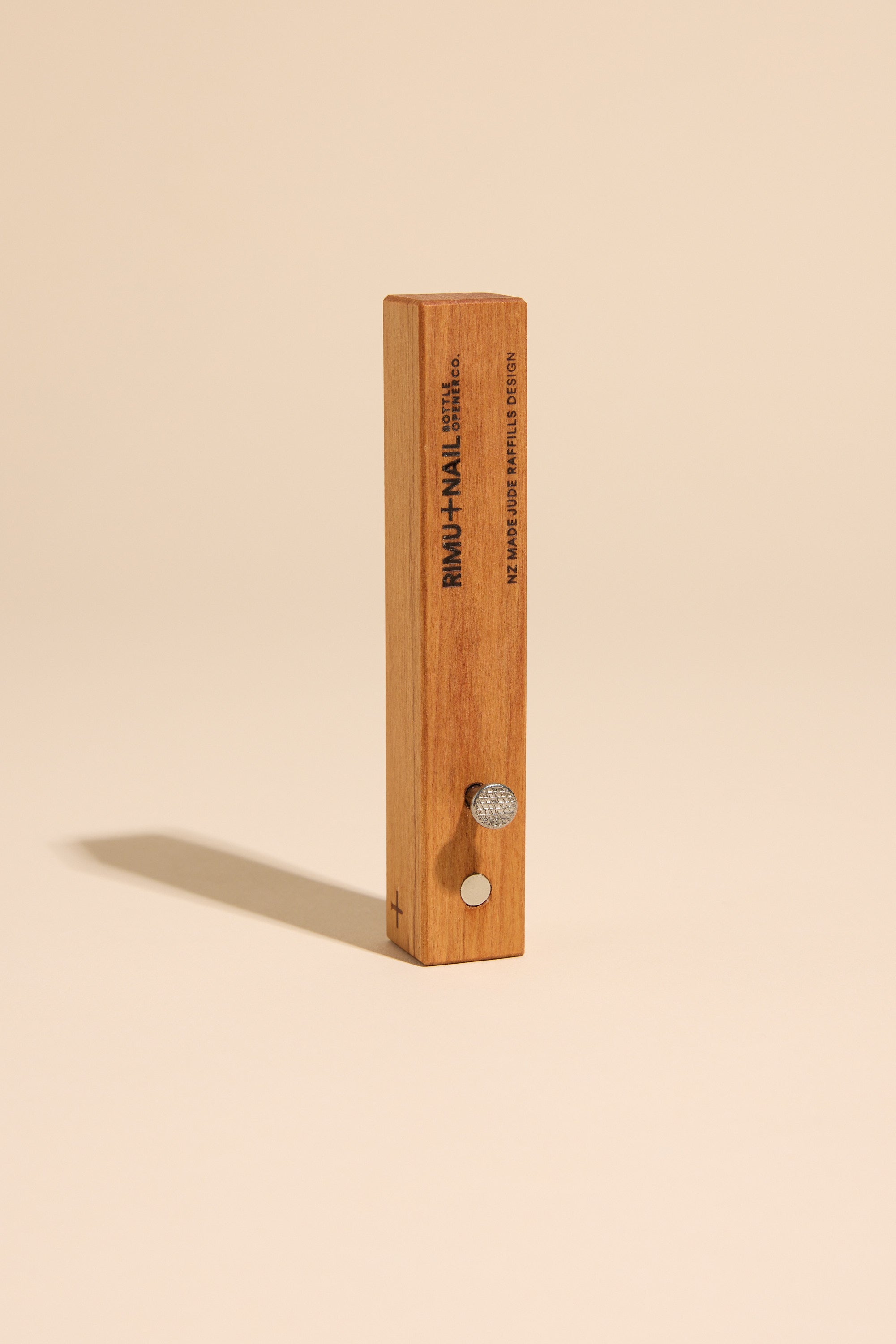The Rimu & Nail Bottle Opener by Other stands upright on a beige surface, featuring a round metal knob, a small button, and engraved details on the front—showcasing its unique, handmade NZ design.