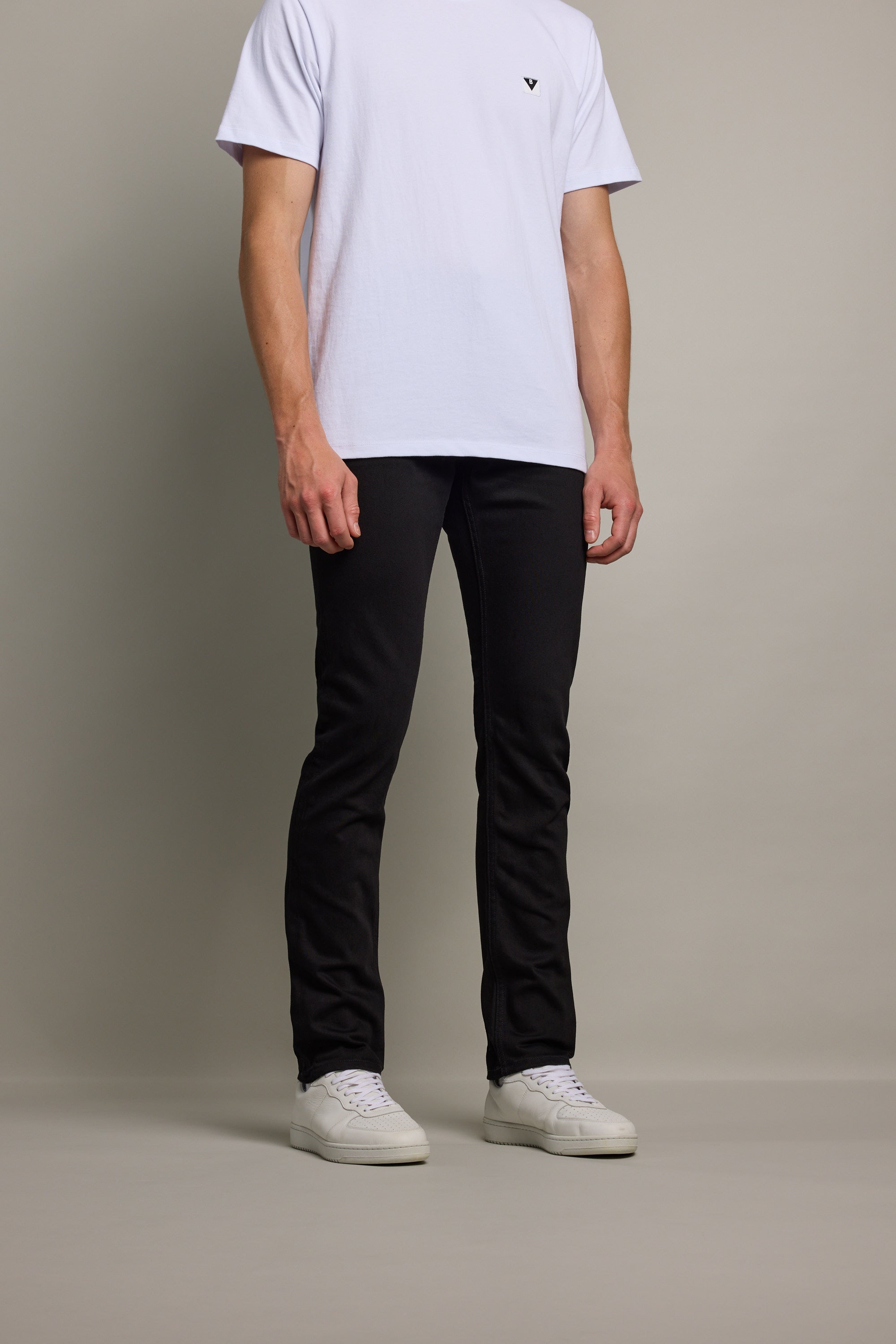 A person, cropped at the shoulders, stands against a plain background wearing Barkers Rambler Denim Slim Jean in black, a white T-shirt, and white sneakers.