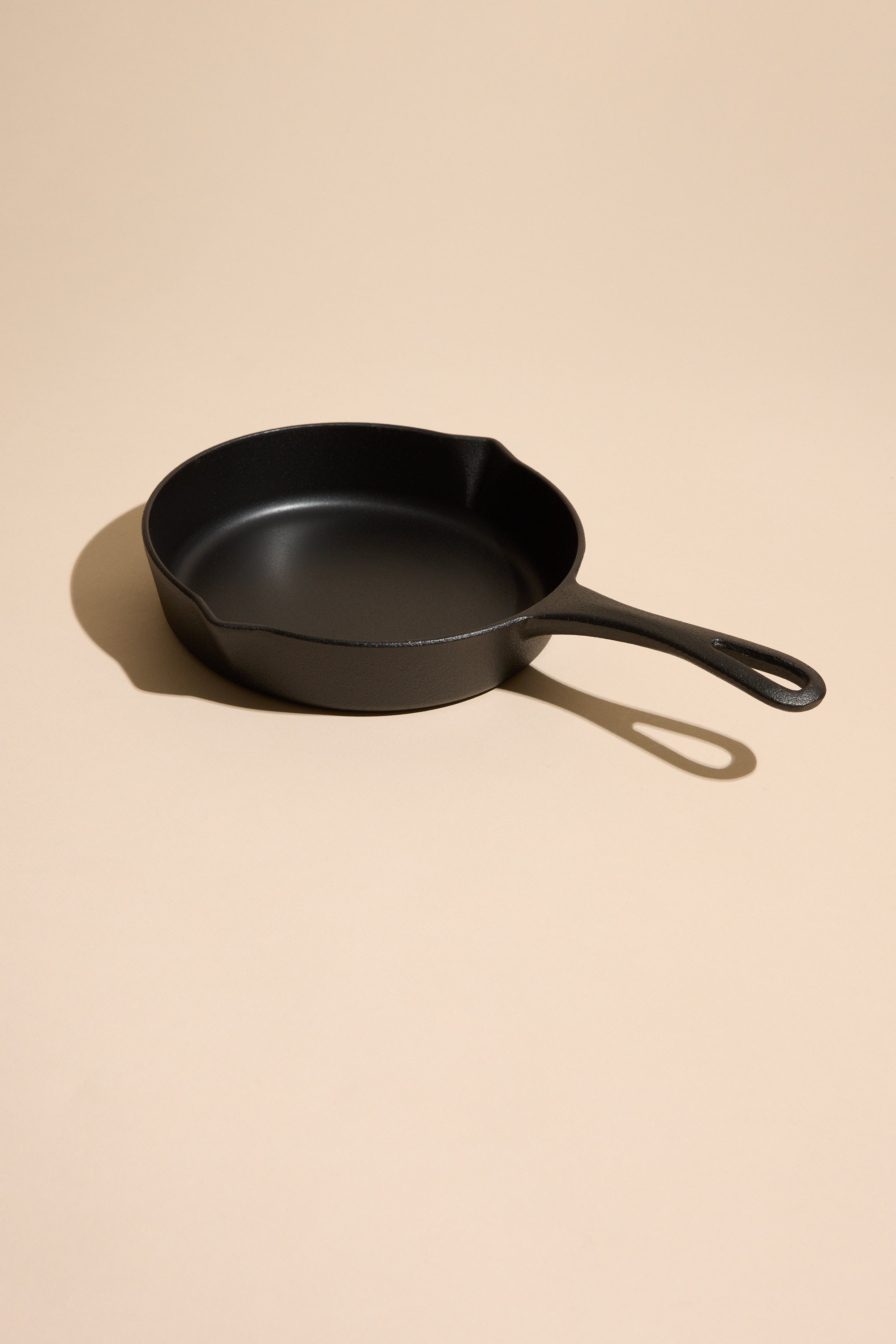 The Ironclad Lil' Legacy Pan by Other, a compact cast iron skillet with a handle, sits on a light beige surface and casts a gentle shadow to the left.