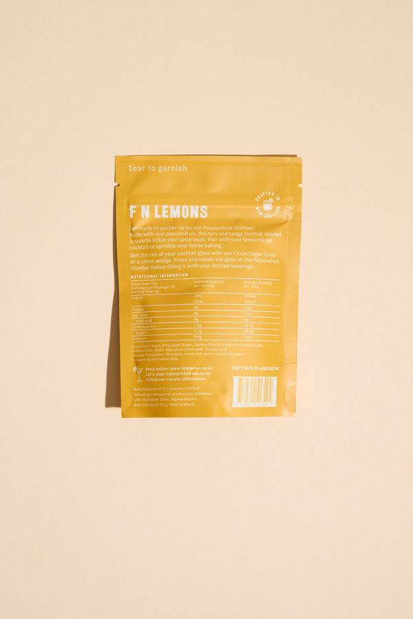 A yellow F N Lemons - Passionfruit Sherbet packet stands upright on a beige background. The back shows white text with product info and nutrition facts. The top reads "Tear to garnish" and "F N LEMONS," ideal as a cocktail garnish or rim dust.