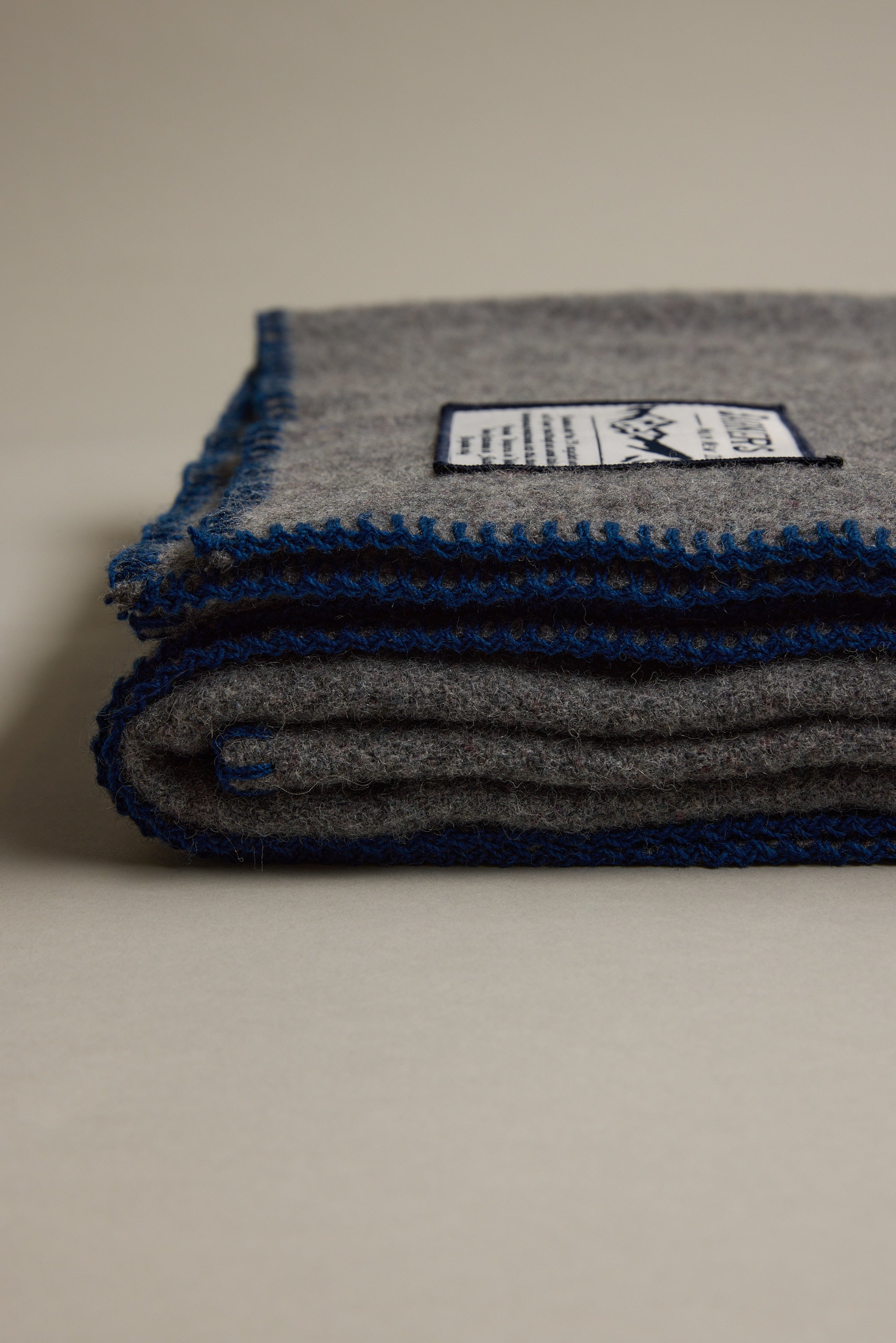 The Wool Take Back Blanket by Other is a folded gray sustainable wool blanket with blue edging and a white label, displayed on a neutral surface.