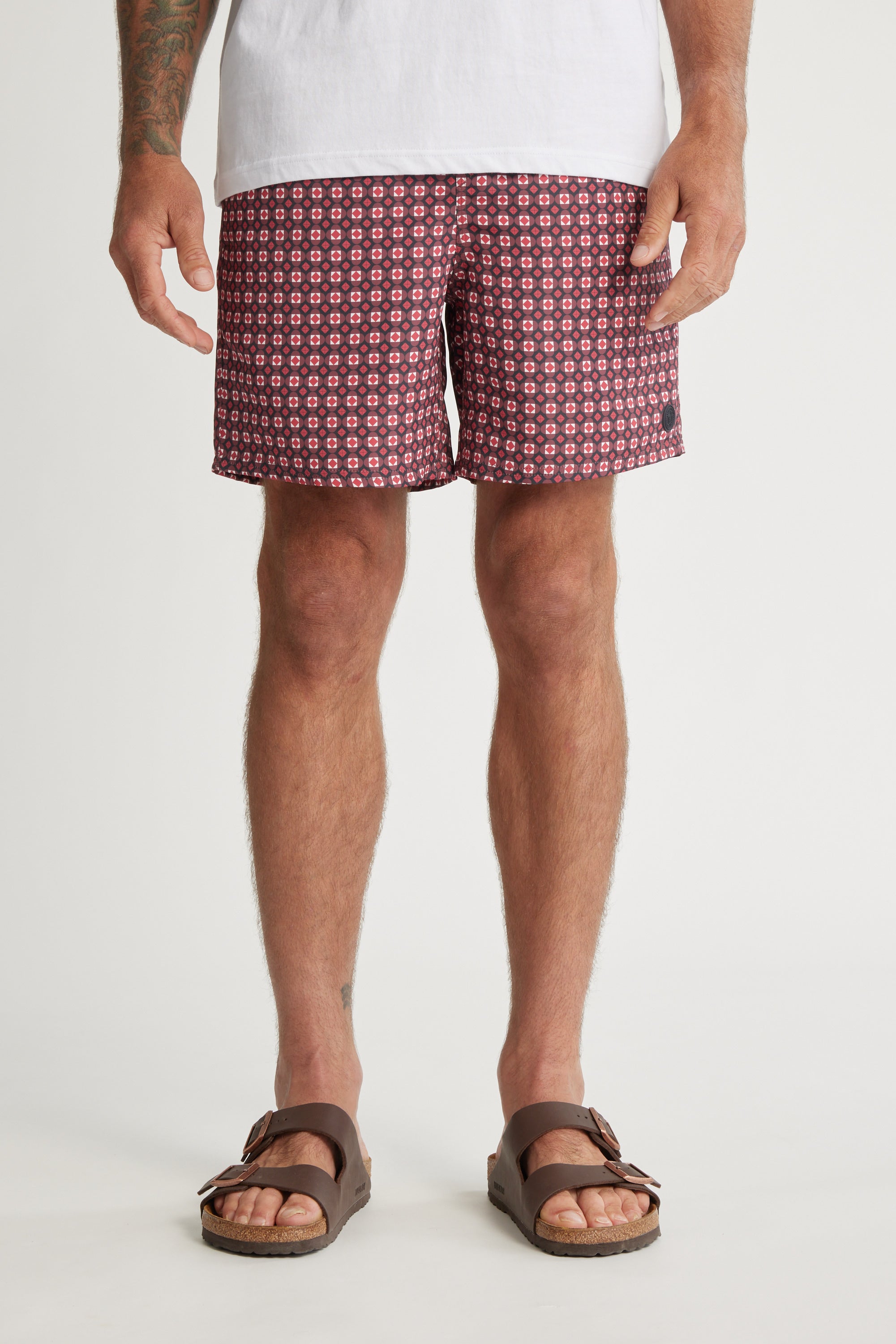 A person wearing a white T-shirt, Barkers Laramie Print Volley shorts with a maroon geometric design made from recycled polyester, and brown sandals stands against a plain white background.