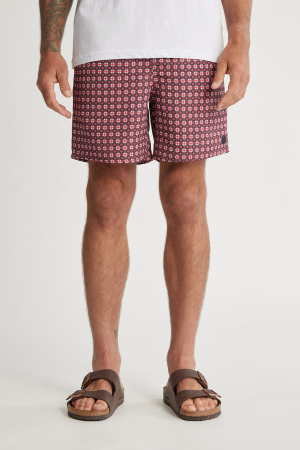 A person wearing a white T-shirt, Barkers Laramie Print Volley shorts with a maroon geometric design made from recycled polyester, and brown sandals stands against a plain white background.