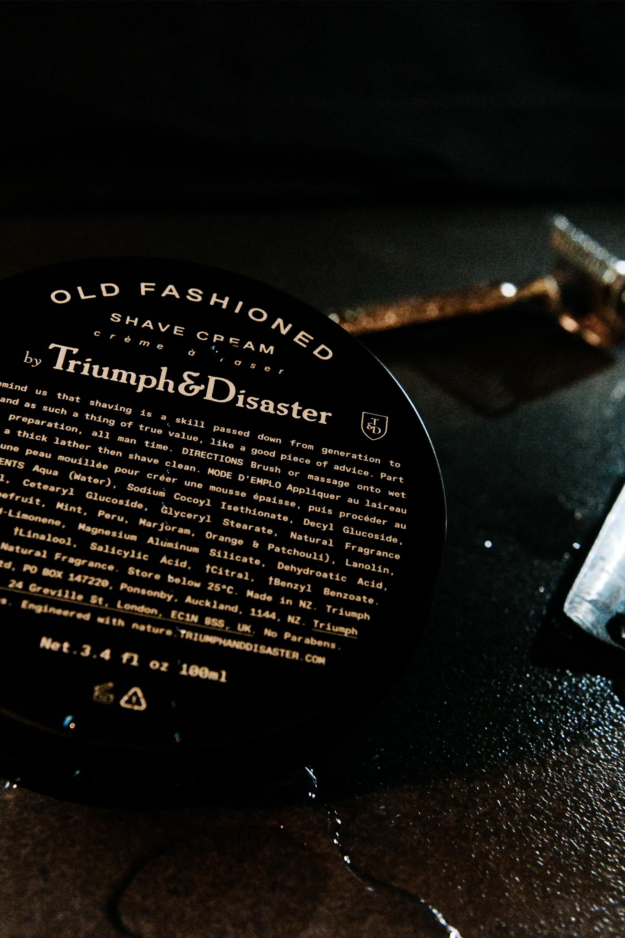A close-up of the Triumph & Disaster Old Fashioned Shave Cream Jar rests on a dark surface, with a gold razor and a glass object softly blurred in the background, capturing traditional shaving style.